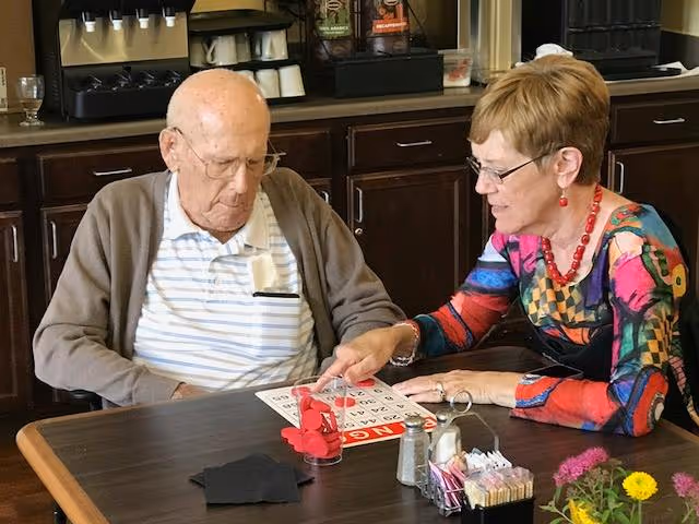 Two seniors sitting at a table in a dining area playing bingo, with a coffee station and cabinets in the background.