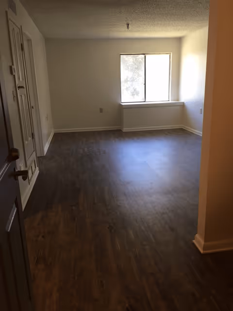 Empty apartment living room with dark wood-look flooring, white walls, and a single window at the far wall.