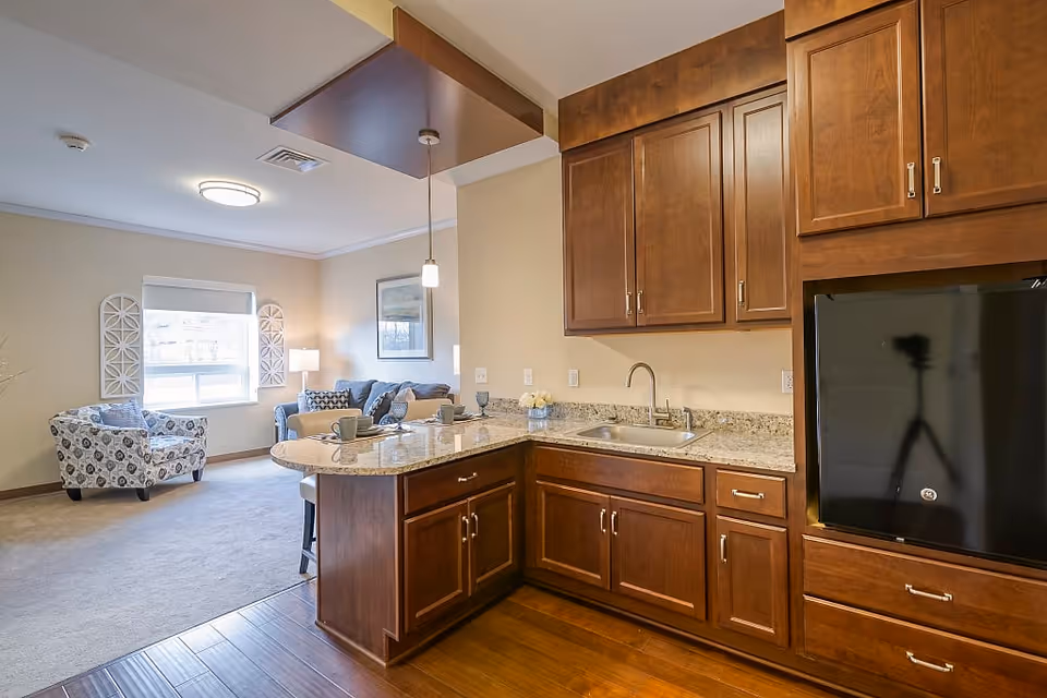 Interior view of a senior living facility showing a kitchen area with wooden cabinets, granite countertops, a sink, and a small refrigerator. Adjacent to the kitchen is a living room area with a patterned armchair, a sofa, a window with a roller shade, and wall decorations. The space is well-lit with ceiling lights and a pendant light above the kitchen counter.