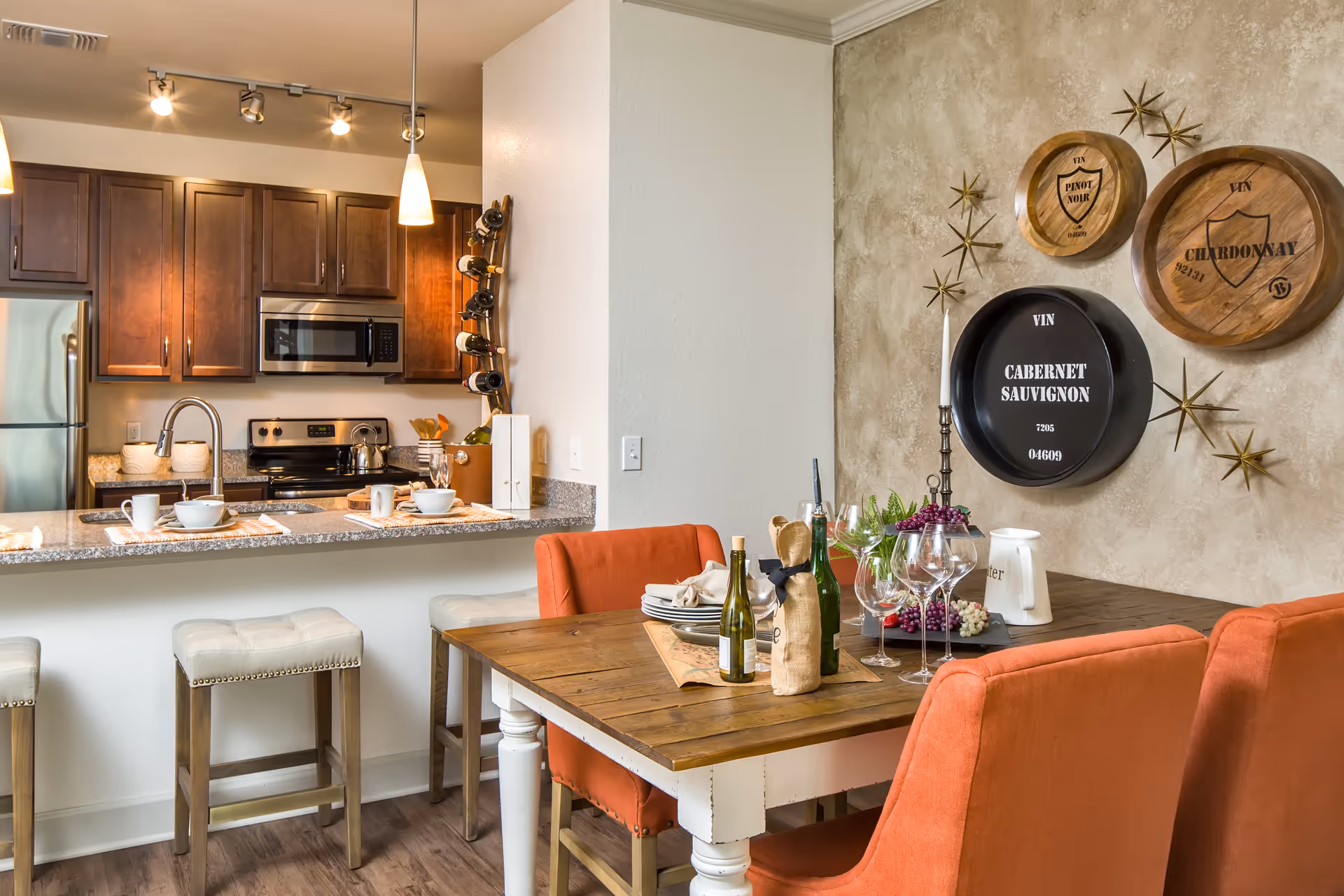Open-plan dining area with a wooden table set for wine and orange upholstered chairs next to a kitchen with granite counter and stainless steel appliances.