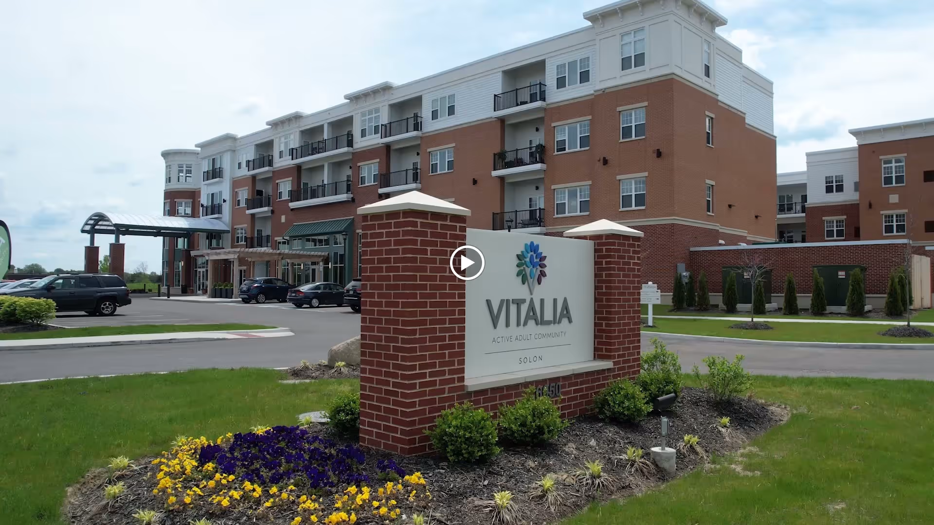 Front exterior view of the Vitalia Active Adult Community at Solon building with a brick sign in the foreground displaying the community name and logo, surrounded by landscaping and parked cars near the entrance.