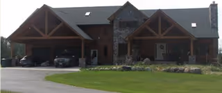 Front exterior view of a large rustic lodge-style building with wooden beams, stone accents, a covered porch, and a driveway with parked vehicles.