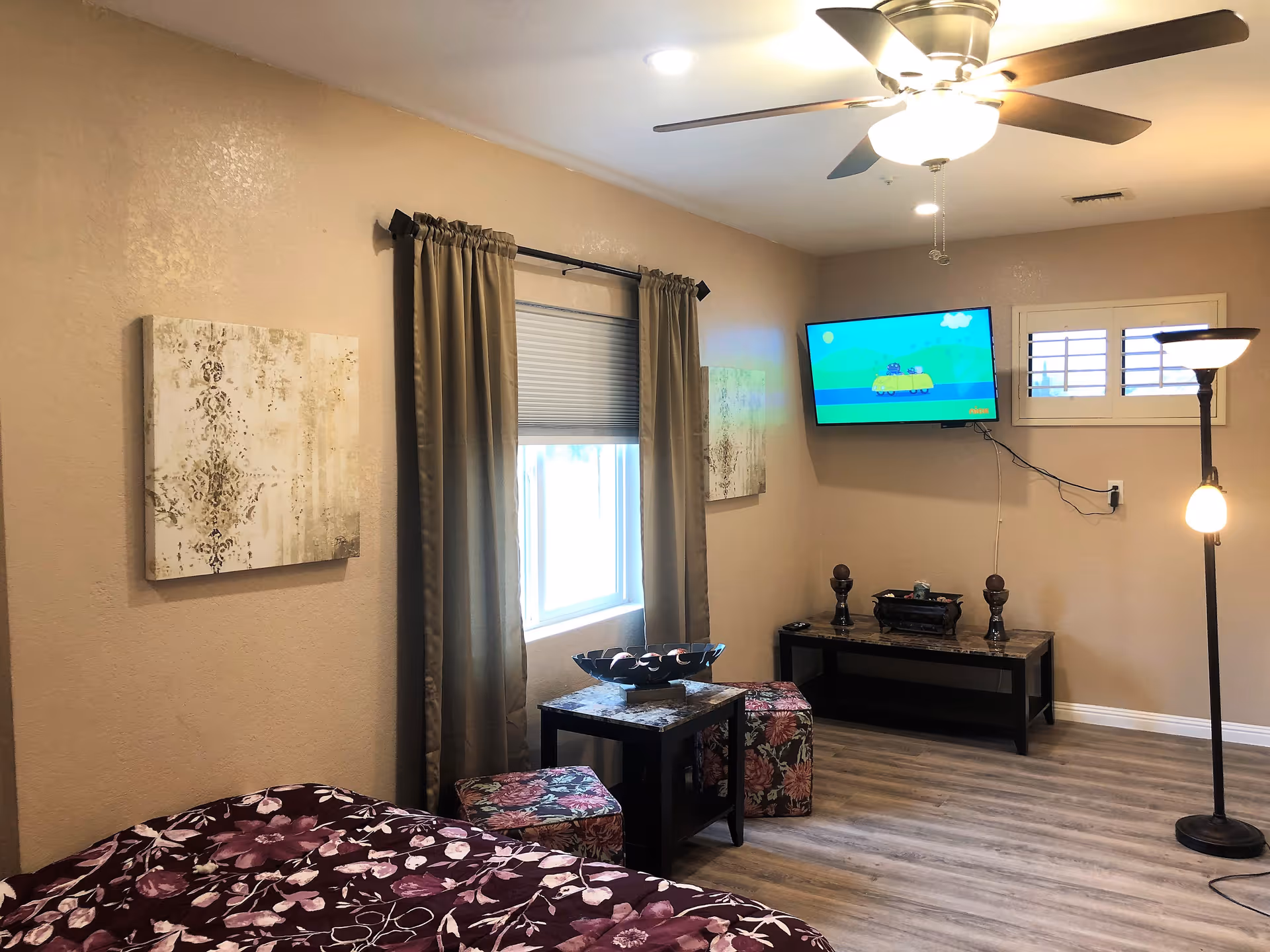 A cozy living room area with beige walls and wood flooring. There is a ceiling fan with lights, a wall-mounted TV showing a cartoon, a floor lamp, a small table with decorative items, and two floral-patterned ottomans. A window with curtains and a blind is on one wall, and a floral-patterned bedspread is partially visible in the foreground.