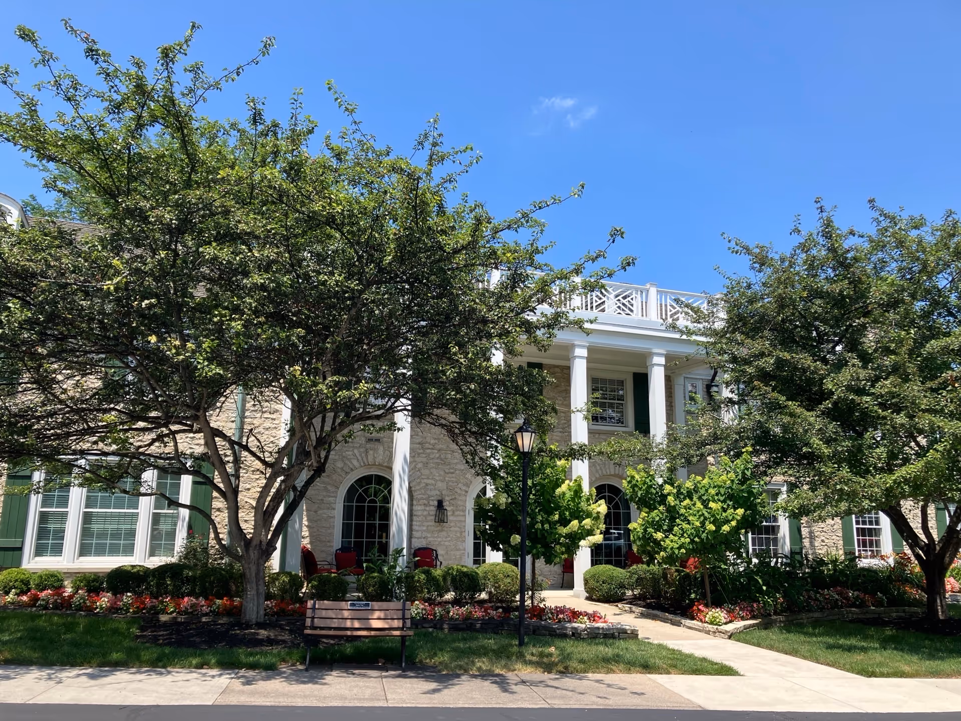 Front exterior view of a senior living facility named Wesley Glen, featuring a stone facade, white columns, green shutters, large windows, and a well-maintained garden with trees, bushes, and flowers under a clear blue sky. A bench and a street lamp are visible near the sidewalk in front of the building.
