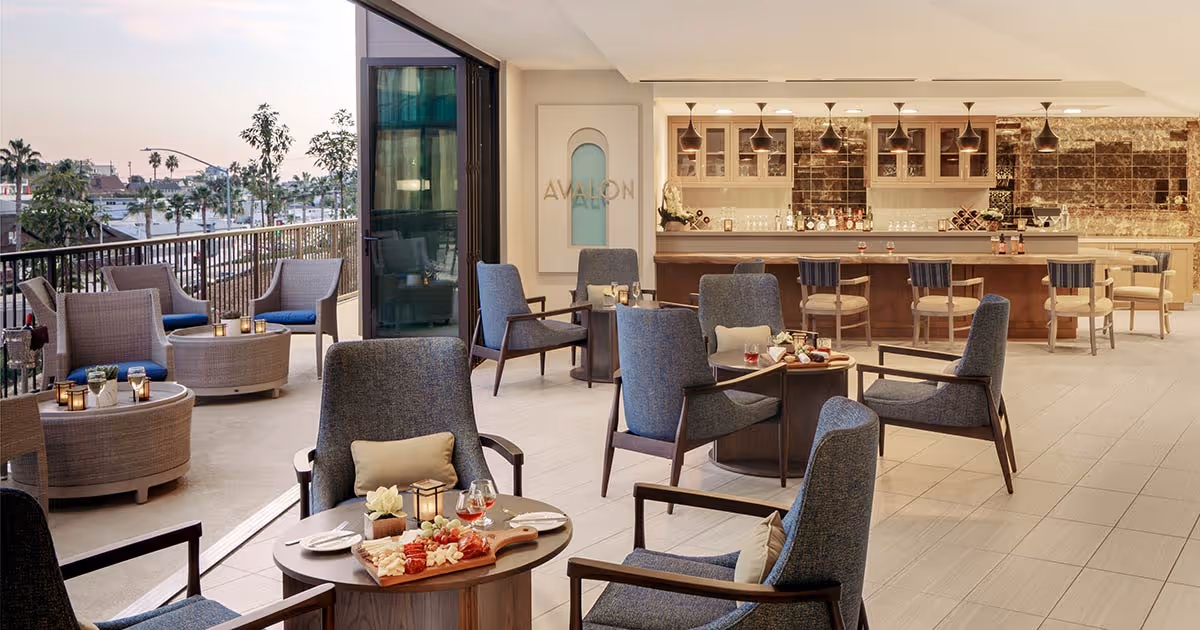 A modern lounge area with comfortable blue armchairs and small round tables set with snacks and drinks. The space opens to an outdoor patio with wicker chairs and tables. In the background, there is a bar with high stools and pendant lighting, and a wall with the word 'AVALON'.
