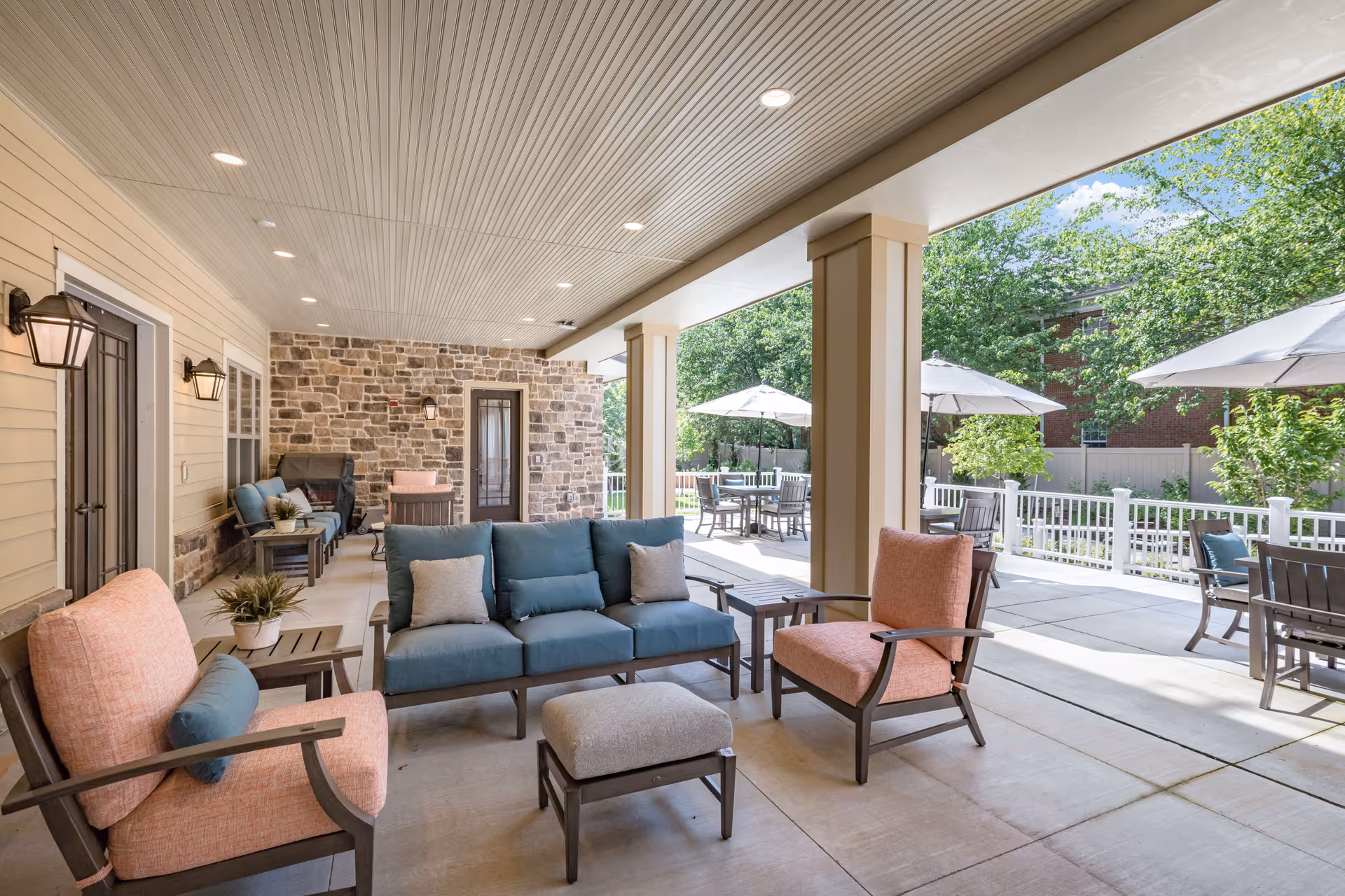 Covered outdoor patio with cushioned sofas and chairs, side tables, and umbrella-shaded dining tables opening onto a sunny courtyard.