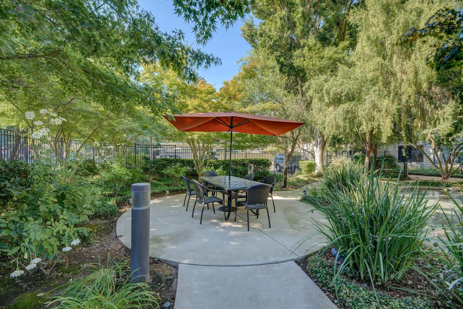 Outdoor patio area with a round concrete seating space featuring a table and six chairs under a large red umbrella, surrounded by lush green trees and plants with a clear blue sky above.