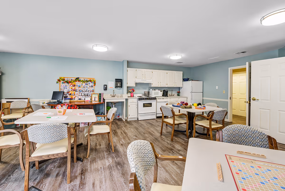 A bright and clean common room with a kitchenette in the background featuring white cabinets, a stove, microwave, and refrigerator. The room has several tables and chairs arranged for group activities, with one table set up for a Scrabble game. The walls are painted light blue, and the floor has wood-style vinyl flooring. There is a bulletin board with photos and decorations on the left wall.