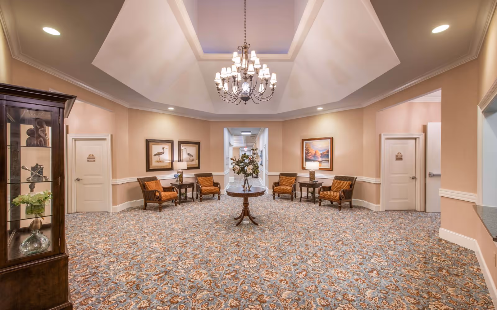 Welcoming senior living facility lobby with a central table of flowers, chandelier, patterned carpet, and seating along the walls.