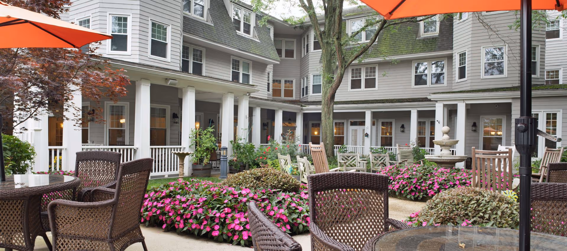 Courtyard patio with wicker tables and chairs, pink flower beds, a fountain, and the multi-story Sunrise at Gardner Park building in the background.