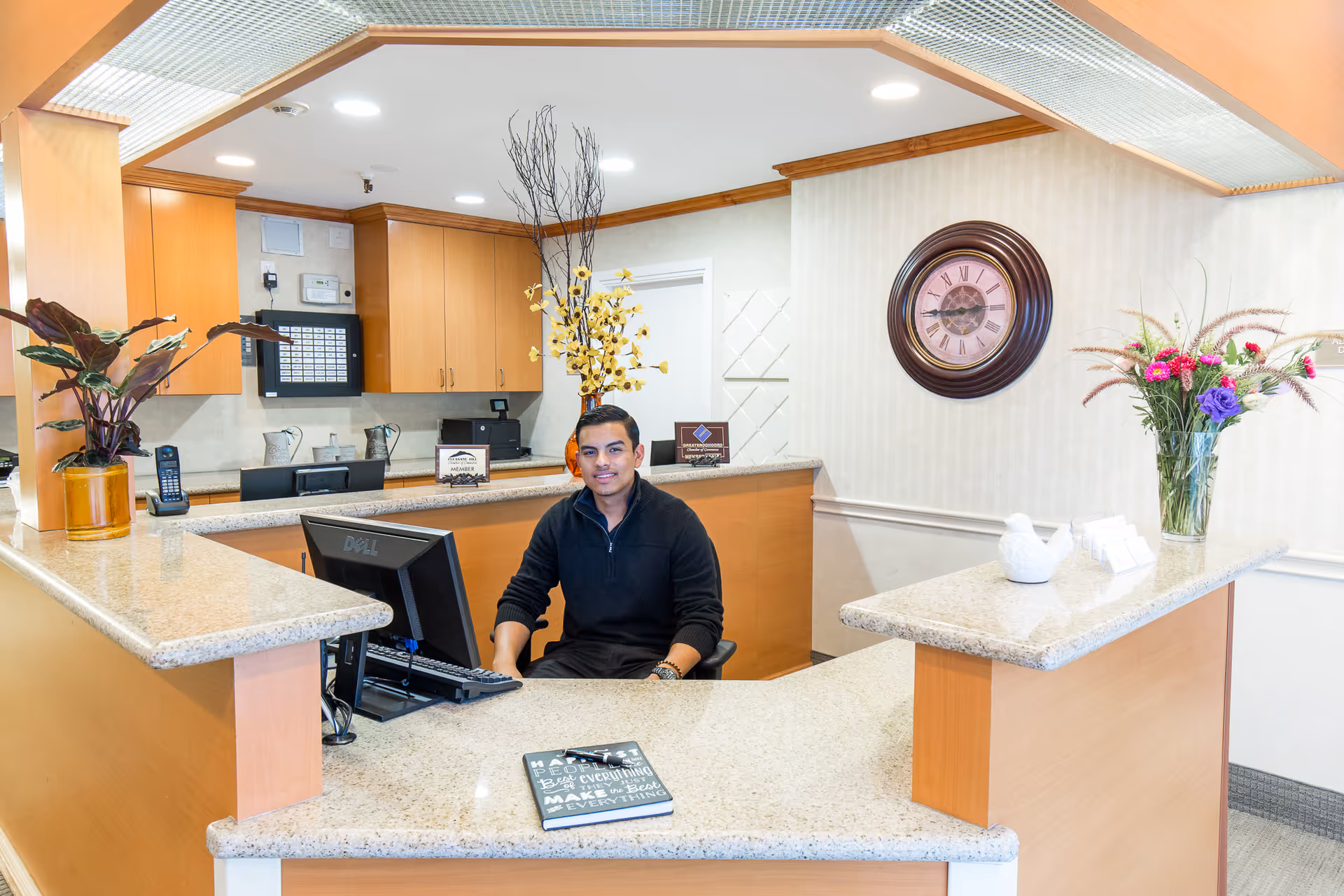 A man sitting behind a reception desk in a well-lit office area with a computer, decorative plants, a wall clock, and a vase with flowers on the counter.