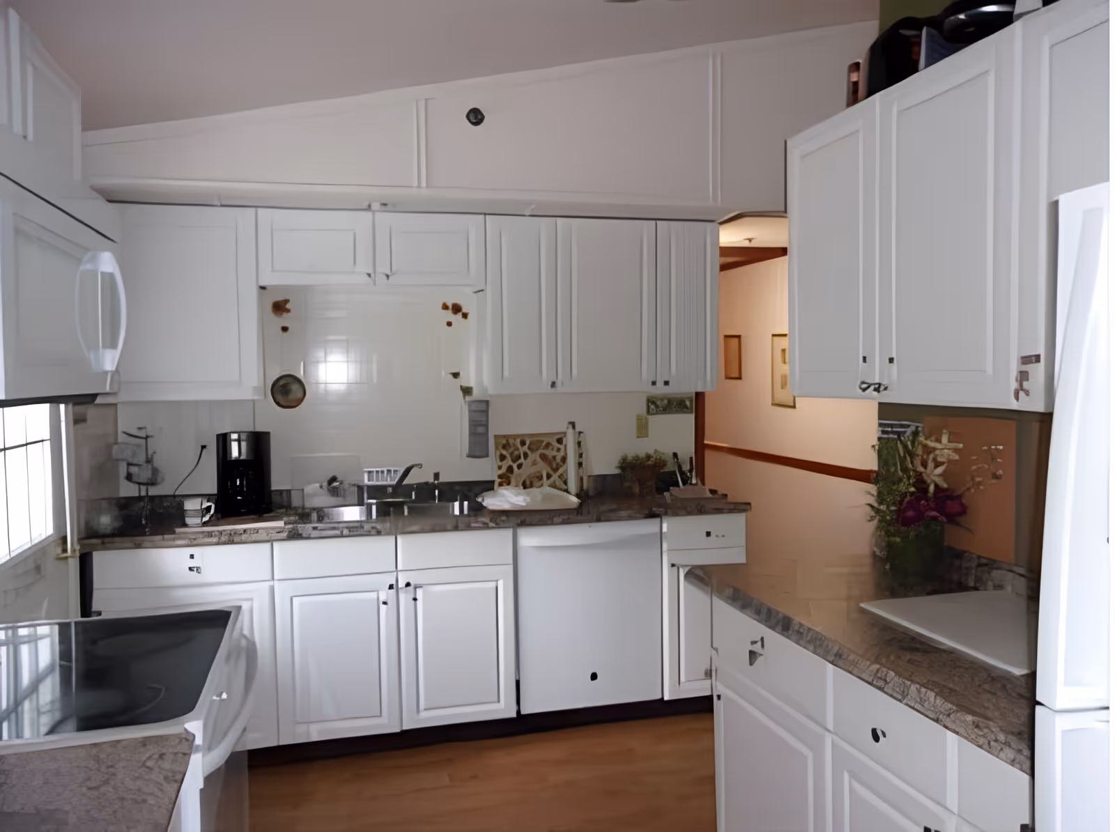 A bright kitchen with white cabinets and drawers, a granite countertop, a black coffee maker, a white dishwasher, and a white refrigerator. There is a window on the left side letting in natural light, and a small vase with flowers on the right countertop. The floor is wooden, and the kitchen opens into a hallway with framed pictures on the wall.