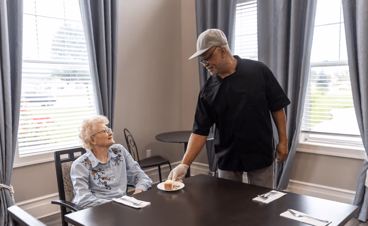 An elderly woman sitting at a dining table in a room with large windows and gray curtains, smiling as a man wearing a black shirt, gray cap, and gloves serves her a small plate with a dessert.