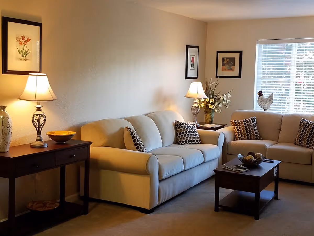 A cozy living room with two beige sofas adorned with patterned throw pillows, a wooden coffee table with decorative items, two side tables each with a lamp, framed floral artwork on the walls, and a window with white blinds letting in natural light.