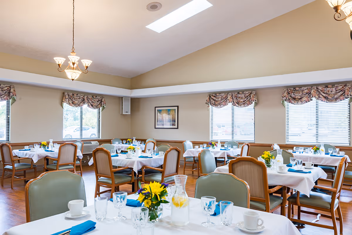 A bright dining room with several tables covered in white tablecloths, each set with glassware, cups, blue napkins, and small flower arrangements. The room has wooden floors, beige walls, multiple windows with floral valances, and a chandelier hanging from the ceiling.