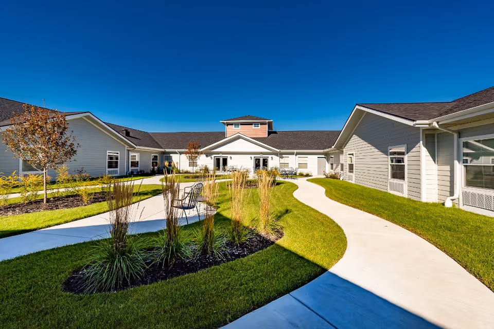 Outdoor courtyard area of a senior living facility with a clear blue sky, green grass, landscaped plants, and a winding concrete pathway. The building surrounds the courtyard with light-colored siding and multiple windows.