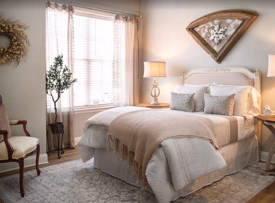 Sunlit cozy bedroom with a made bed, bedside lamps, decorative wall art, and a window with sheer curtains.