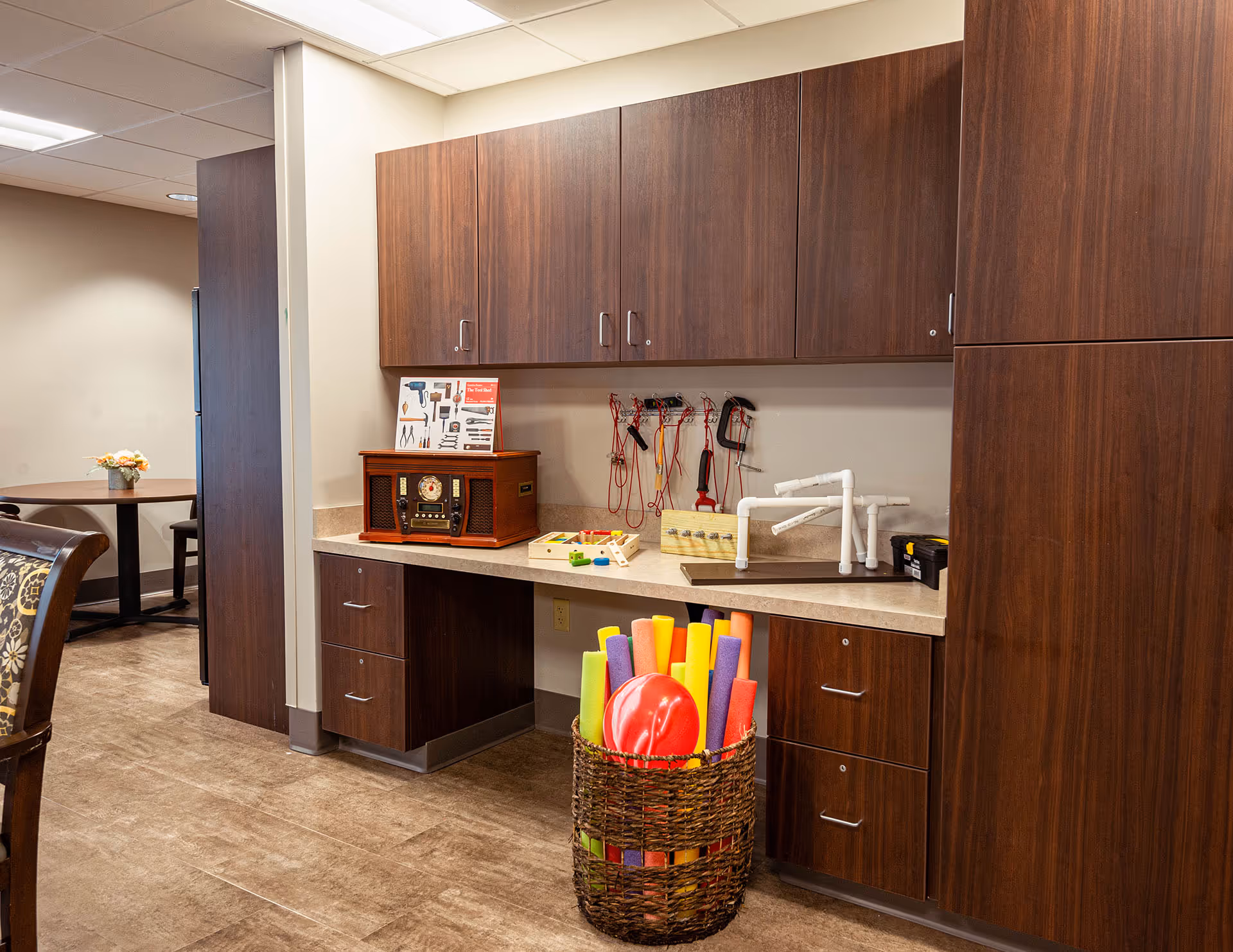 A room with wooden cabinets and a countertop holding various tools and craft items. On the countertop, there is a vintage-style radio, a small wooden pegboard with colorful pegs, and some white PVC pipe structures. Hanging on the wall above the counter are several hand tools with red handles. In front of the counter, there is a woven basket filled with colorful pool noodles and a red ball. To the left, there is a partial view of a dining area with a round table and chairs, and a small flower arrangement on the table.