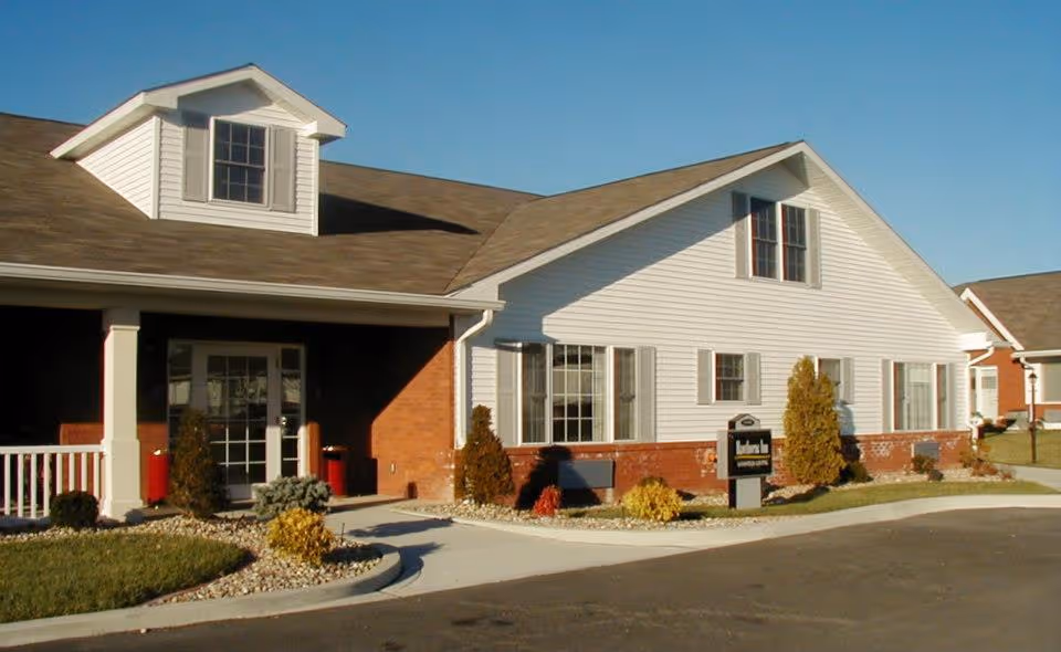 Exterior view of a single-story building with white siding and brick accents, featuring multiple windows and a small porch area with white railing. The building is surrounded by a landscaped area with shrubs and a paved driveway under a clear blue sky.