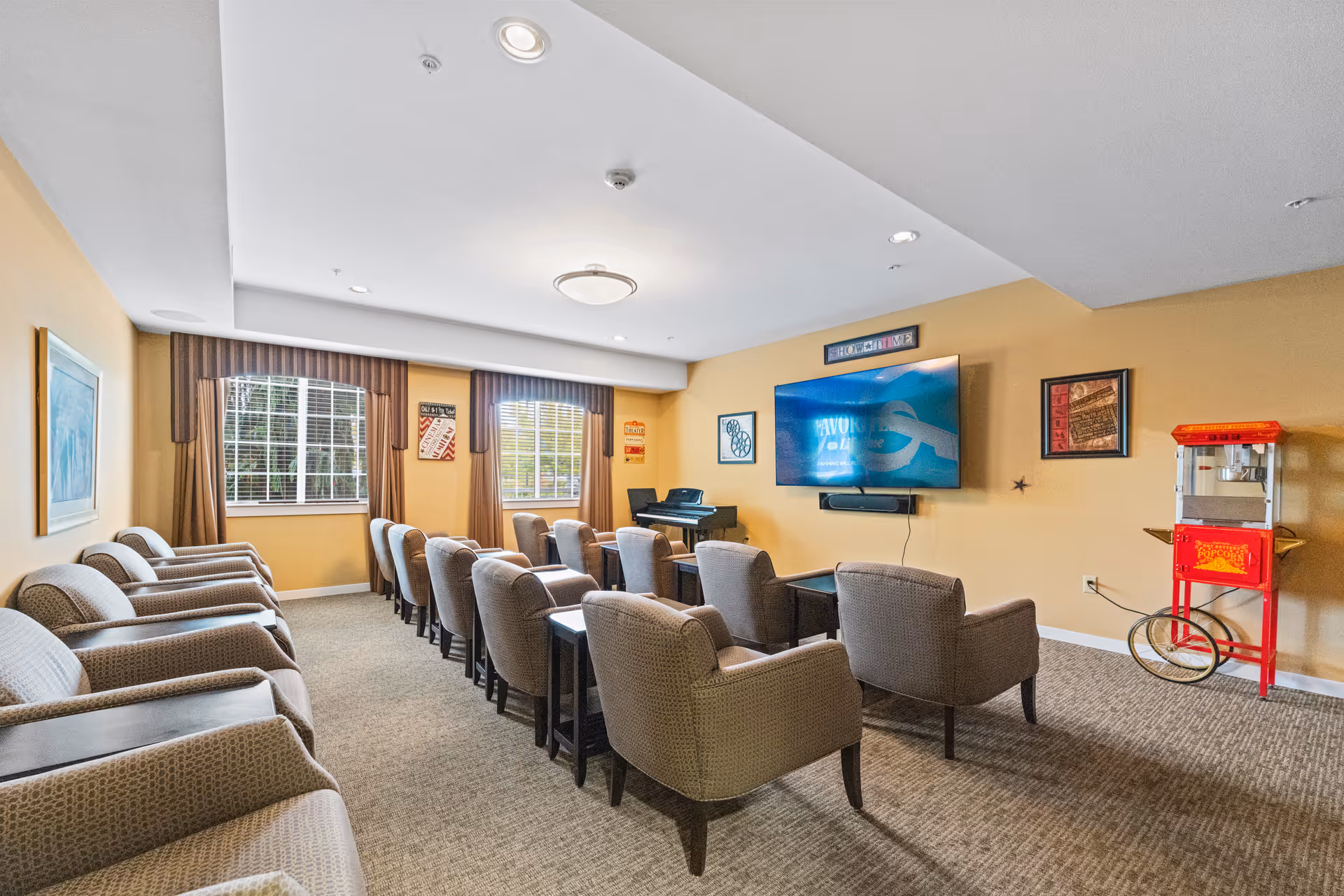 A senior living facility common room with rows of cushioned armchairs facing a large wall-mounted flat screen TV. The room has beige walls, carpeted floor, two windows with brown curtains, framed artwork on the walls, a piano in the corner, and a vintage-style red popcorn machine on wheels.