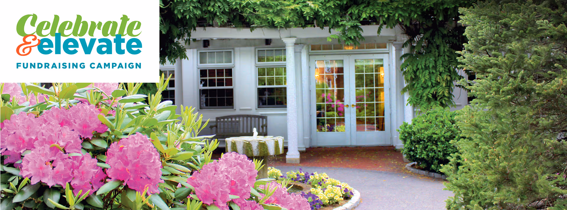 Entrance to a building surrounded by lush greenery and blooming pink flowers, with a pathway leading to double glass doors framed by white columns. A bench and a small fountain are visible near the entrance.