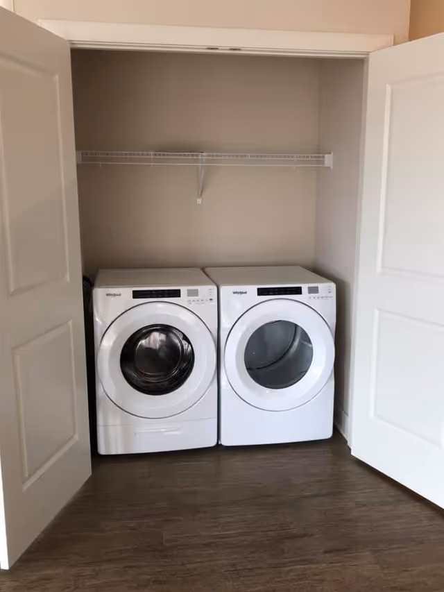 Two front-loading washer and dryer units side by side inside a closet with open doors and a wire shelf above.