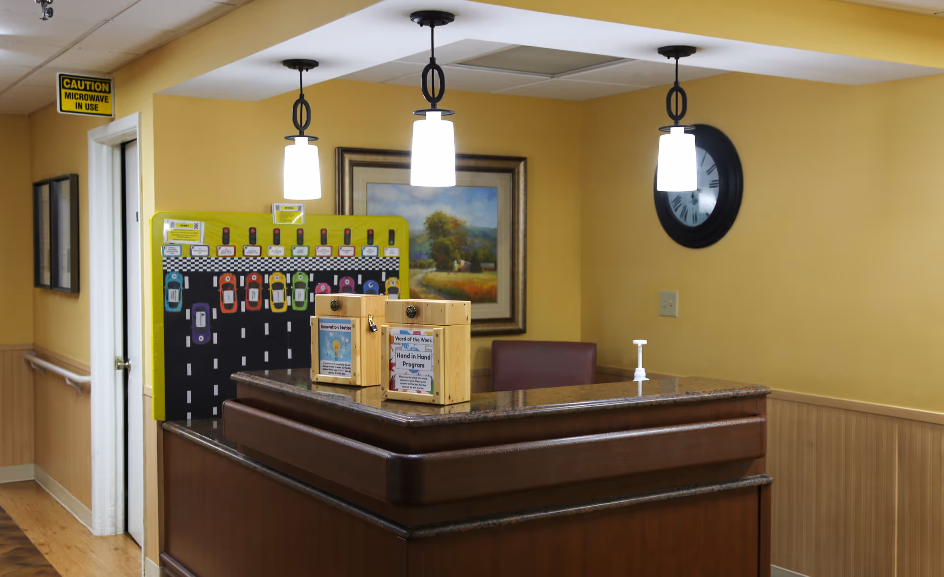 Reception desk area with a dark wooden counter and three hanging pendant lights above. Behind the desk is a framed landscape painting and a large black wall clock. On the desk are two wooden boxes with informational signs and a hand sanitizer dispenser. The walls are painted yellow with wood paneling on the lower half. A door with a sign above it reading 'CAUTION MICROWAVE IN USE' is visible to the left.