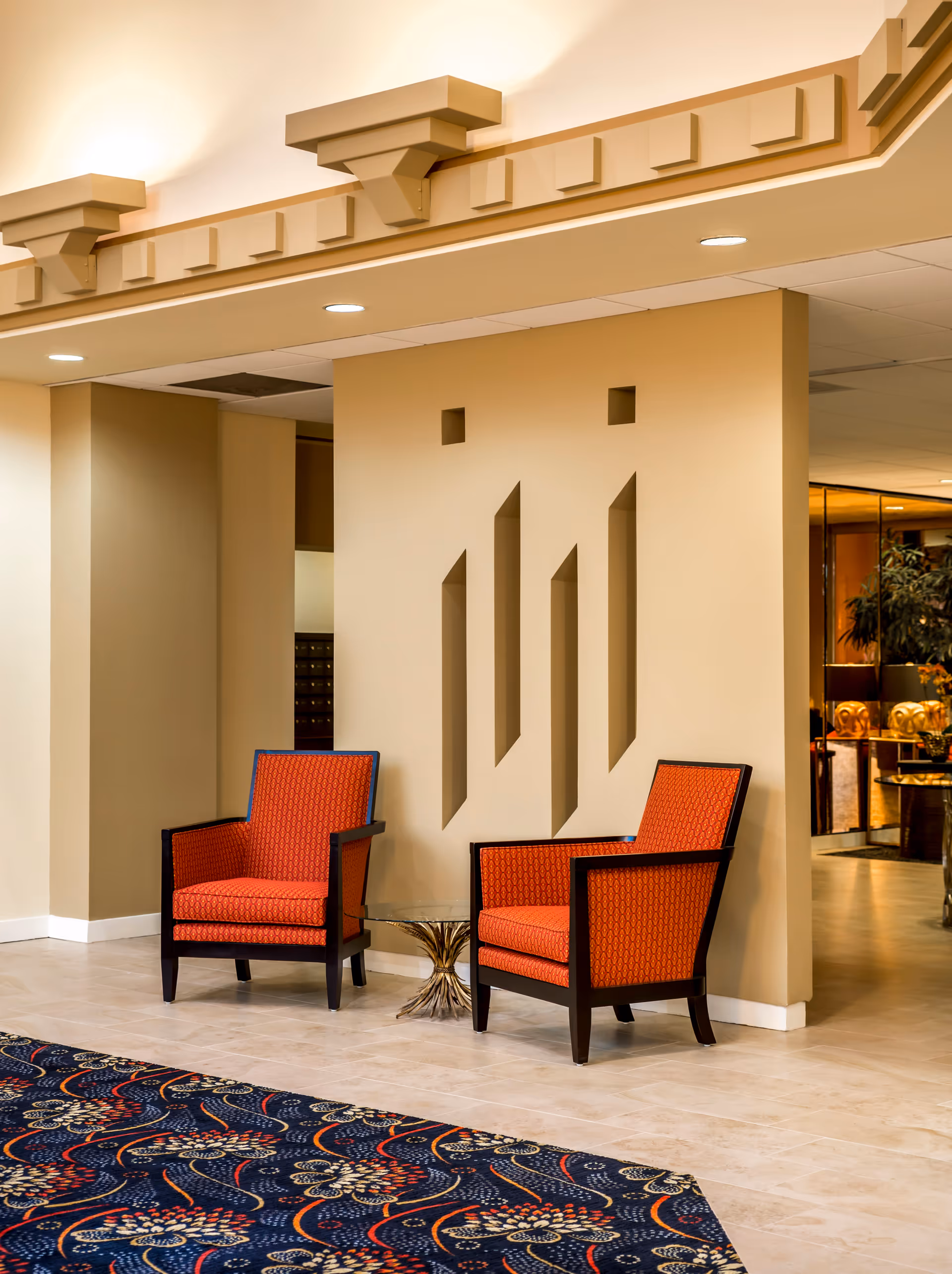 Two orange upholstered armchairs with dark wooden frames are positioned around a small glass-top table with a gold base in a well-lit interior space. The wall behind the chairs features geometric cut-out designs, and the floor is tiled with a patterned carpet partially visible in the foreground.