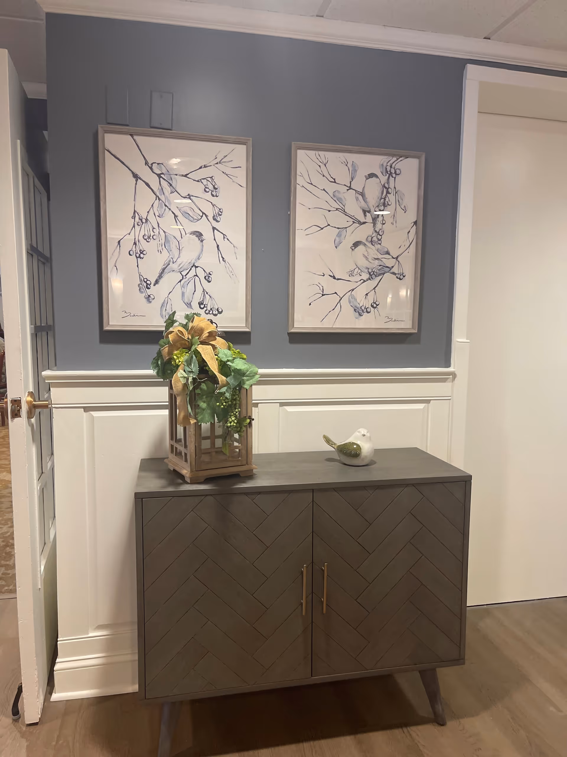 A small wooden cabinet with a herringbone pattern on the doors, topped with a decorative lantern holding green foliage and a small white bird figurine. Above the cabinet, two framed artworks depict birds perched on branches with berries, set against a gray wall with white wainscoting below.