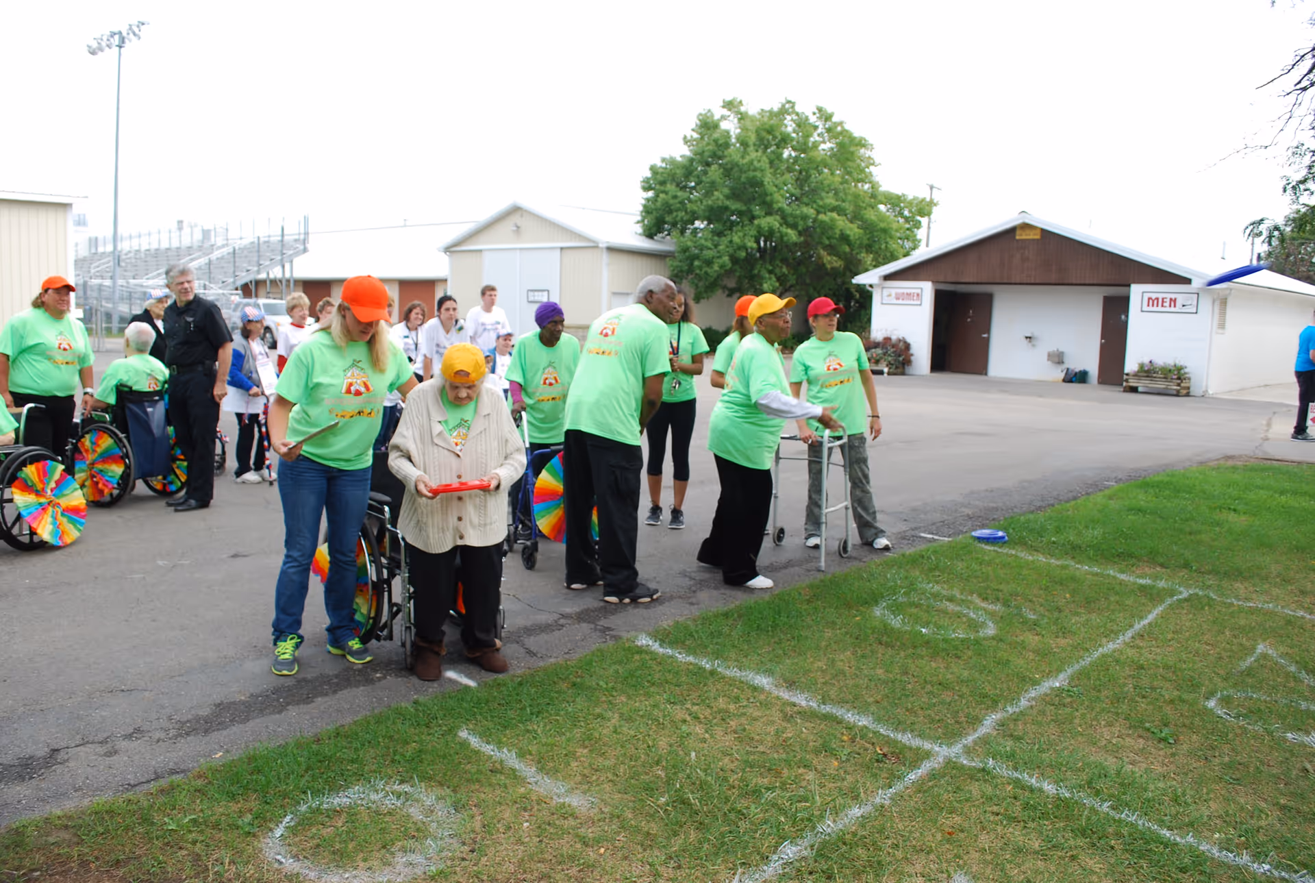 A group of elderly people and caregivers, some in wheelchairs, gathered outdoors on a paved area next to a grassy field with a large tic-tac-toe game marked on the grass. The caregivers and some participants are wearing bright green shirts and colorful hats. In the background, there are buildings with signs indicating restrooms for men and women.