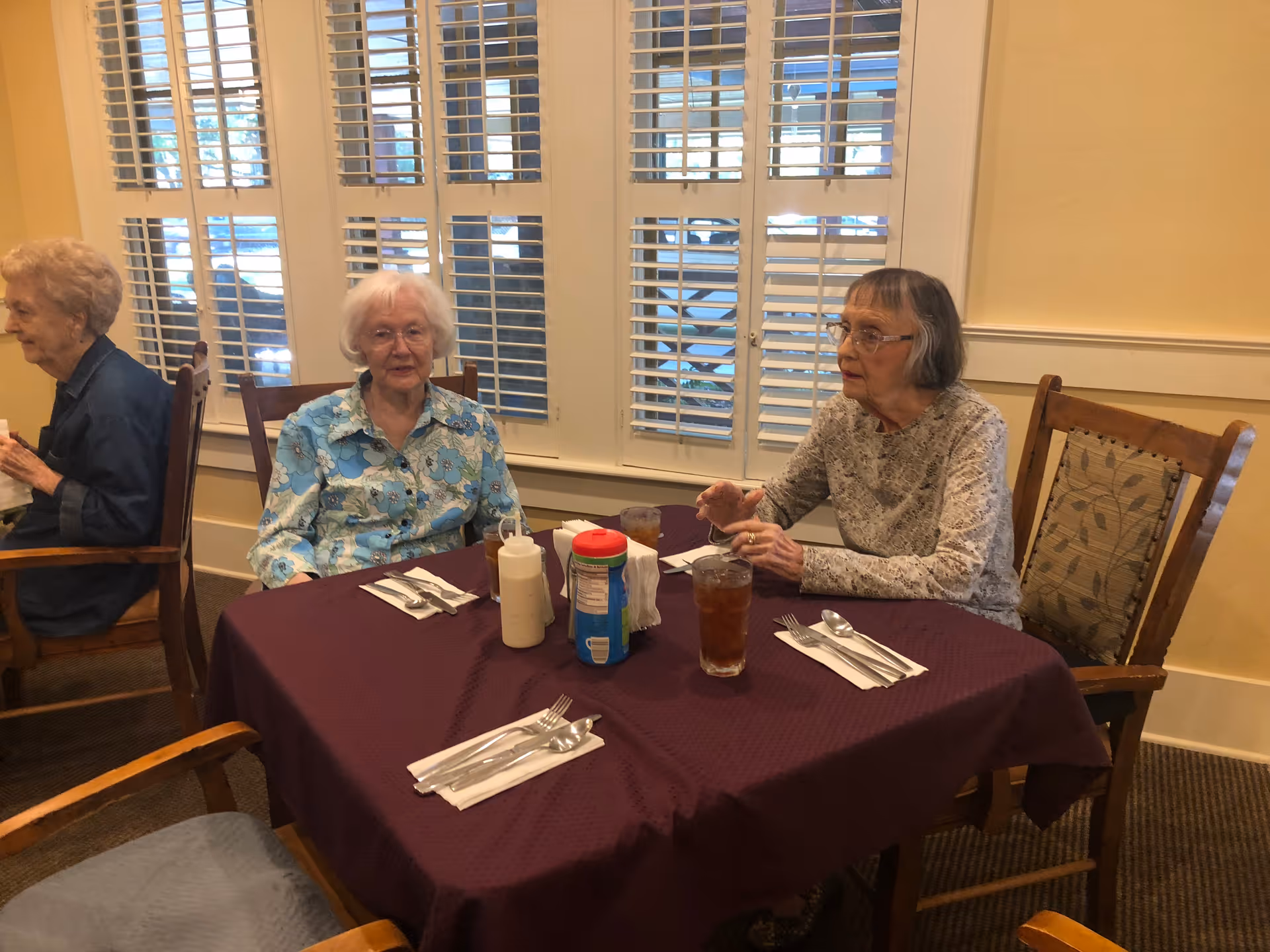 Three older women sit at a table with drinks and condiments in a dining room with shuttered windows.