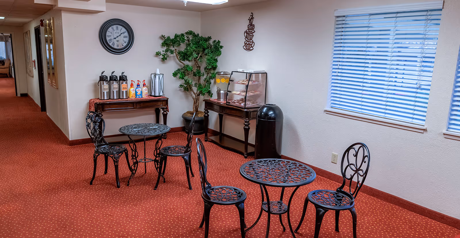 A small seating area in a hallway with two black metal tables and four matching chairs on a red carpet. Against the wall, there are two wooden tables; one holds coffee dispensers, creamers, and cups, while the other has a beverage dispenser with lemon water and a display case with pastries. A large clock and a decorative plant are on the wall, and a window with blinds is on the right side.
