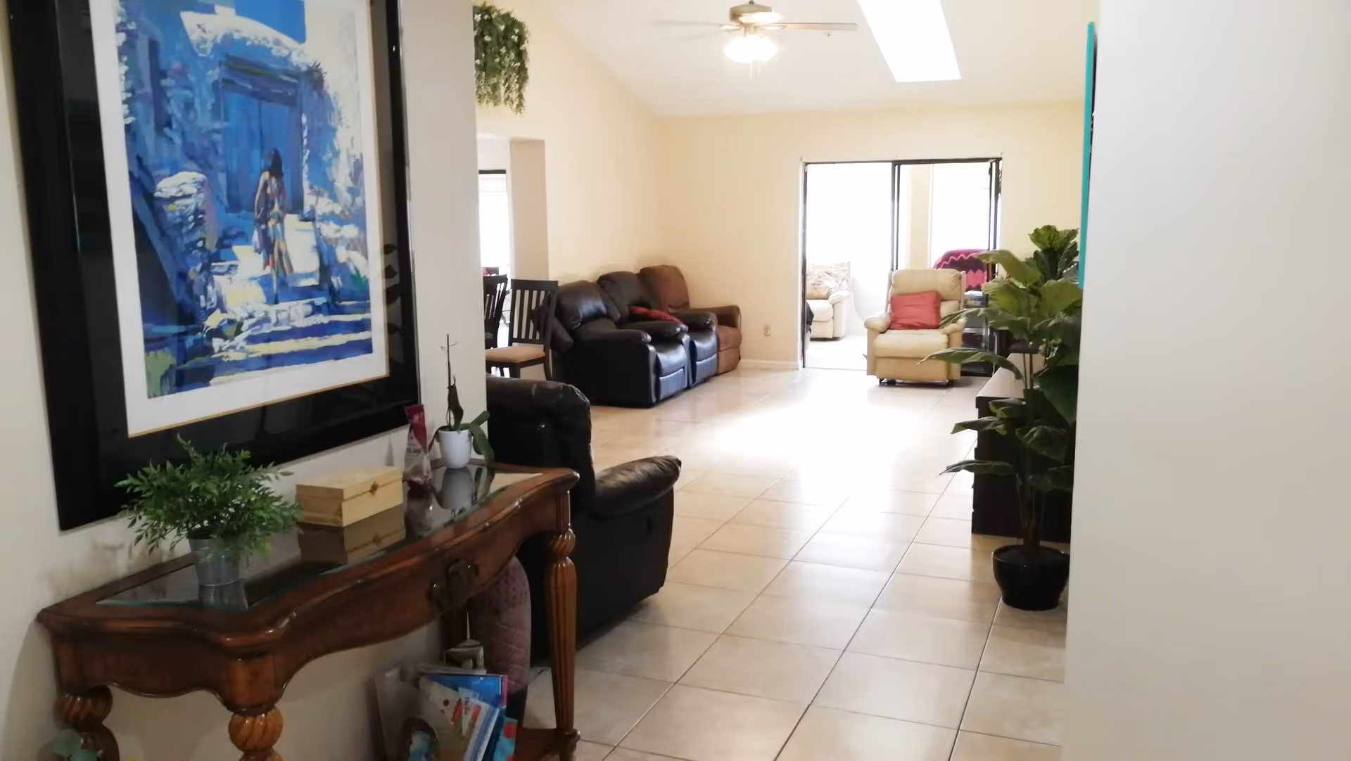 Interior view of a living room area with tiled floors, a ceiling fan, and multiple seating options including leather sofas and armchairs. A wooden console table with plants and decorative items is positioned against the left wall beneath a large framed blue-toned painting. There are plants placed near the right wall and natural light coming through windows in the background.