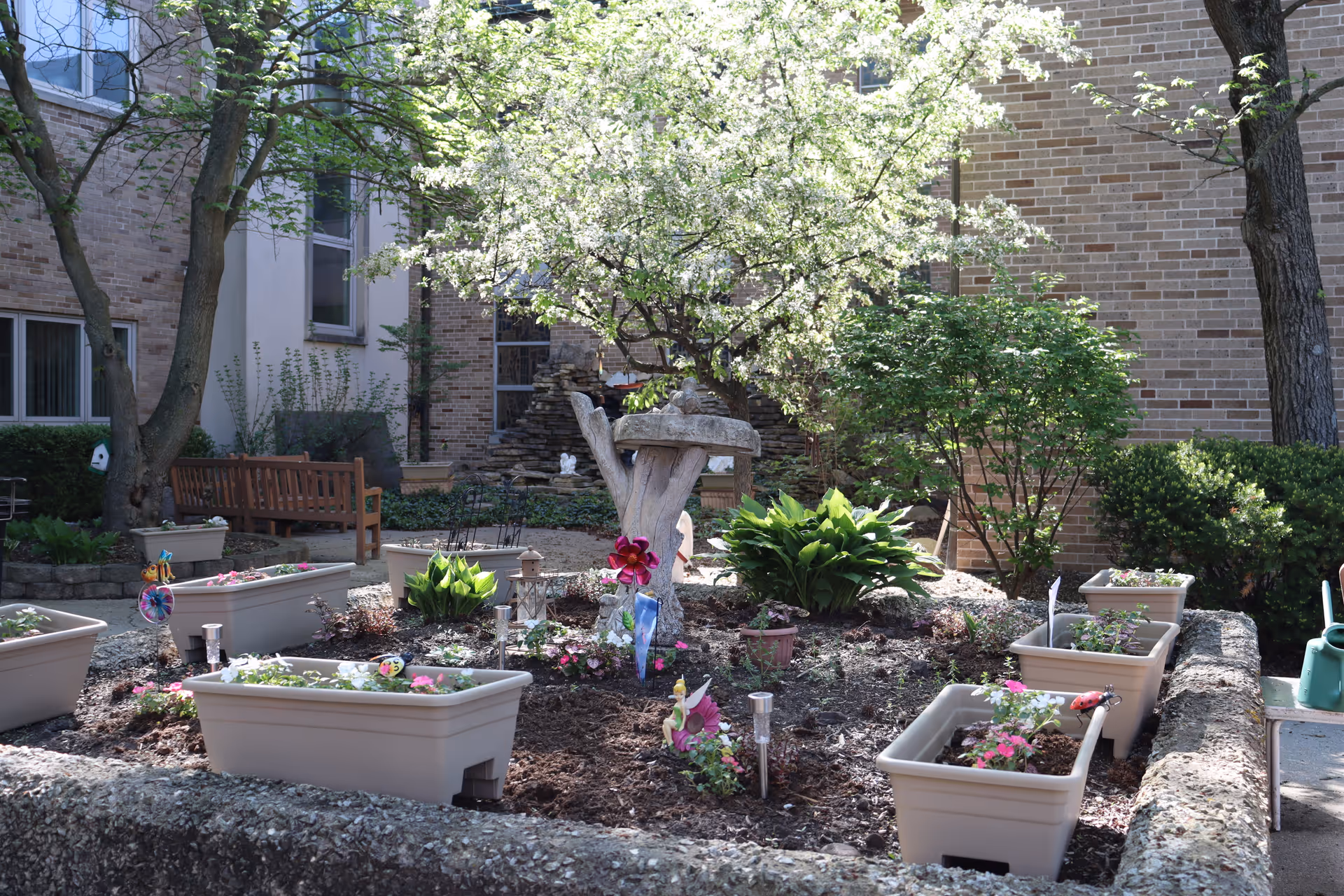Courtyard garden with planter boxes, a birdbath sculpture, benches, and trees outside a brick building.