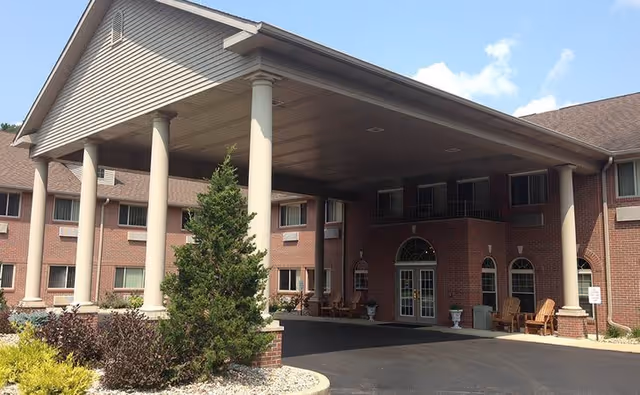 Exterior view of Brentwood at Niles Senior Living facility showing a brick building with multiple windows, a covered entrance supported by large columns, outdoor seating, and landscaped greenery under a partly cloudy sky.