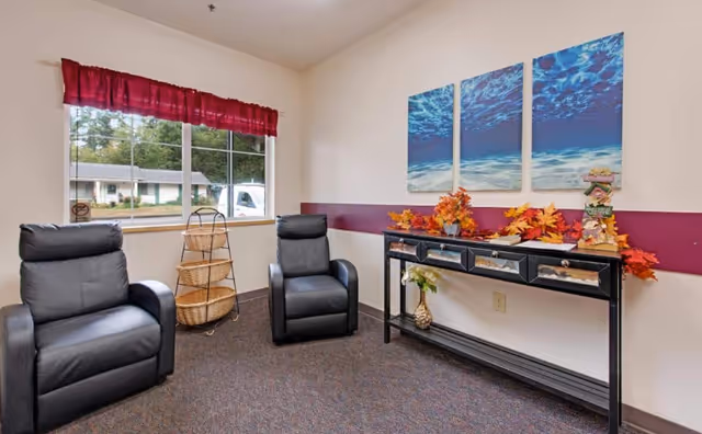 Small seating area with two black recliners by a window and a console table decorated with autumn leaves beneath a three-panel ocean artwork.