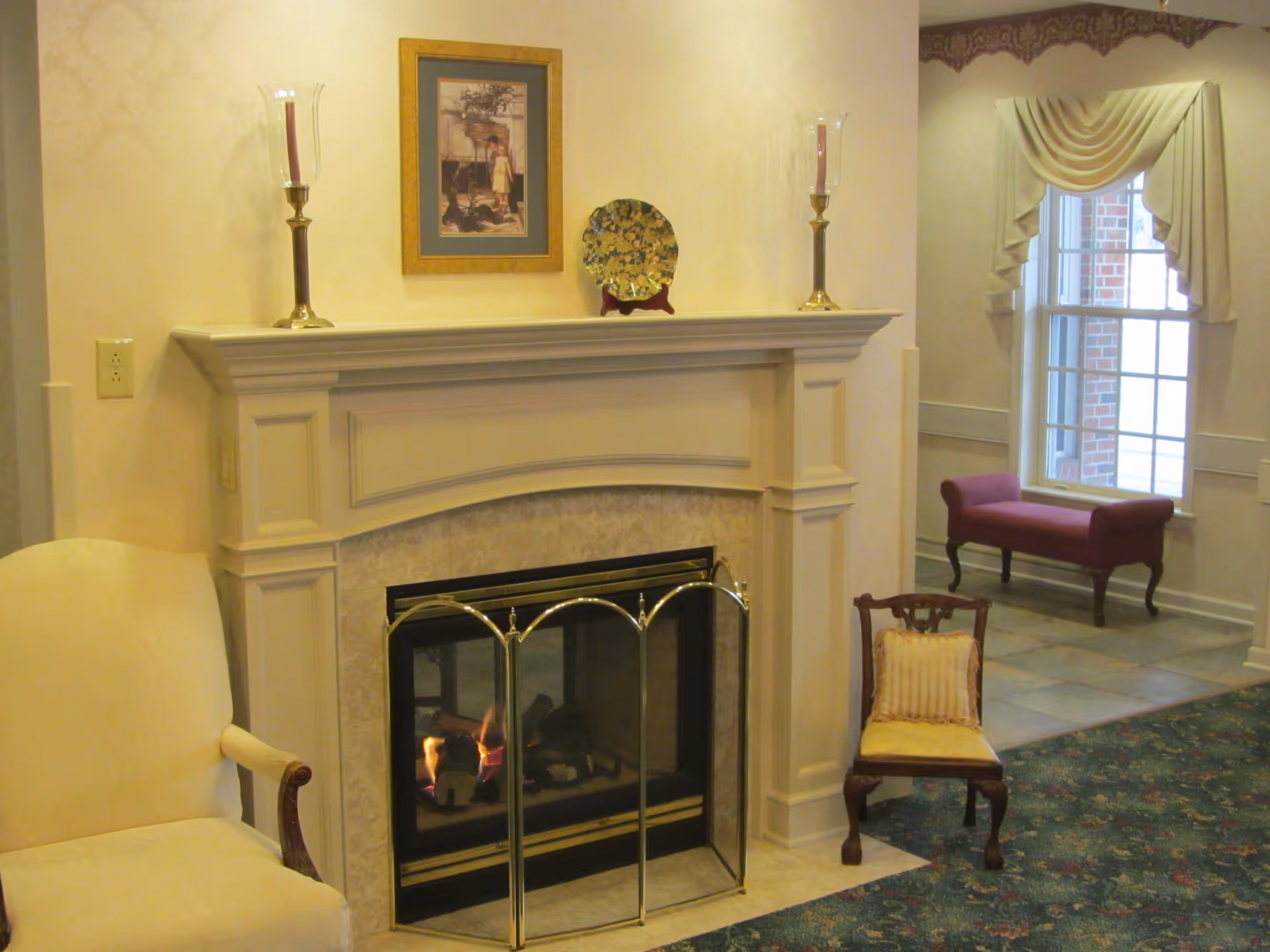 A cozy indoor seating area featuring a lit fireplace with a decorative screen. Above the fireplace mantel are two tall candle holders with candles and a framed picture along with a decorative plate. To the left of the fireplace is a cream-colored armchair, and to the right is a small wooden chair with a cushion. In the background, there is a window with draped curtains and a red upholstered bench beneath it.