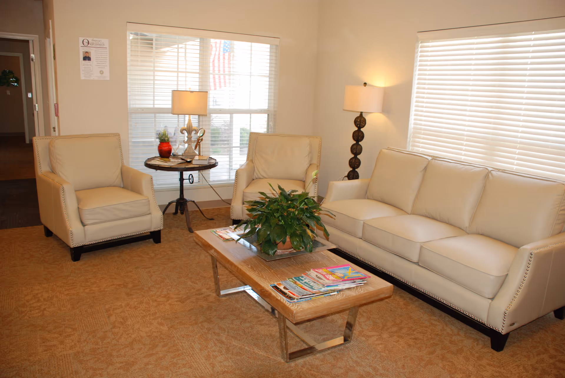 A cozy living room area with a beige three-seater sofa and two matching armchairs. A wooden coffee table with a green potted plant and several magazines is placed in the center. There are two lamps, one on a round side table between the armchairs and another floor lamp near the sofa. Large windows with white blinds allow natural light to fill the room.