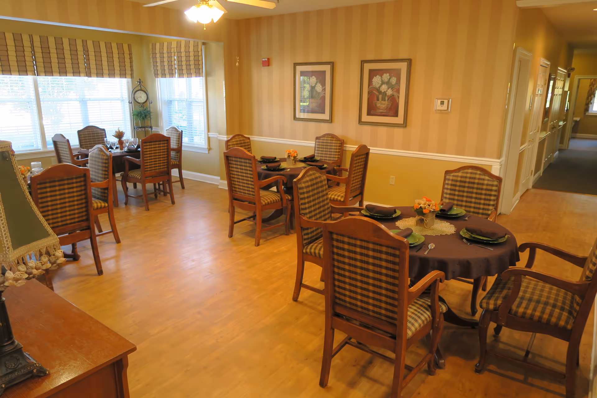 A sunny dining room with round tables set for meals and wooden upholstered chairs.