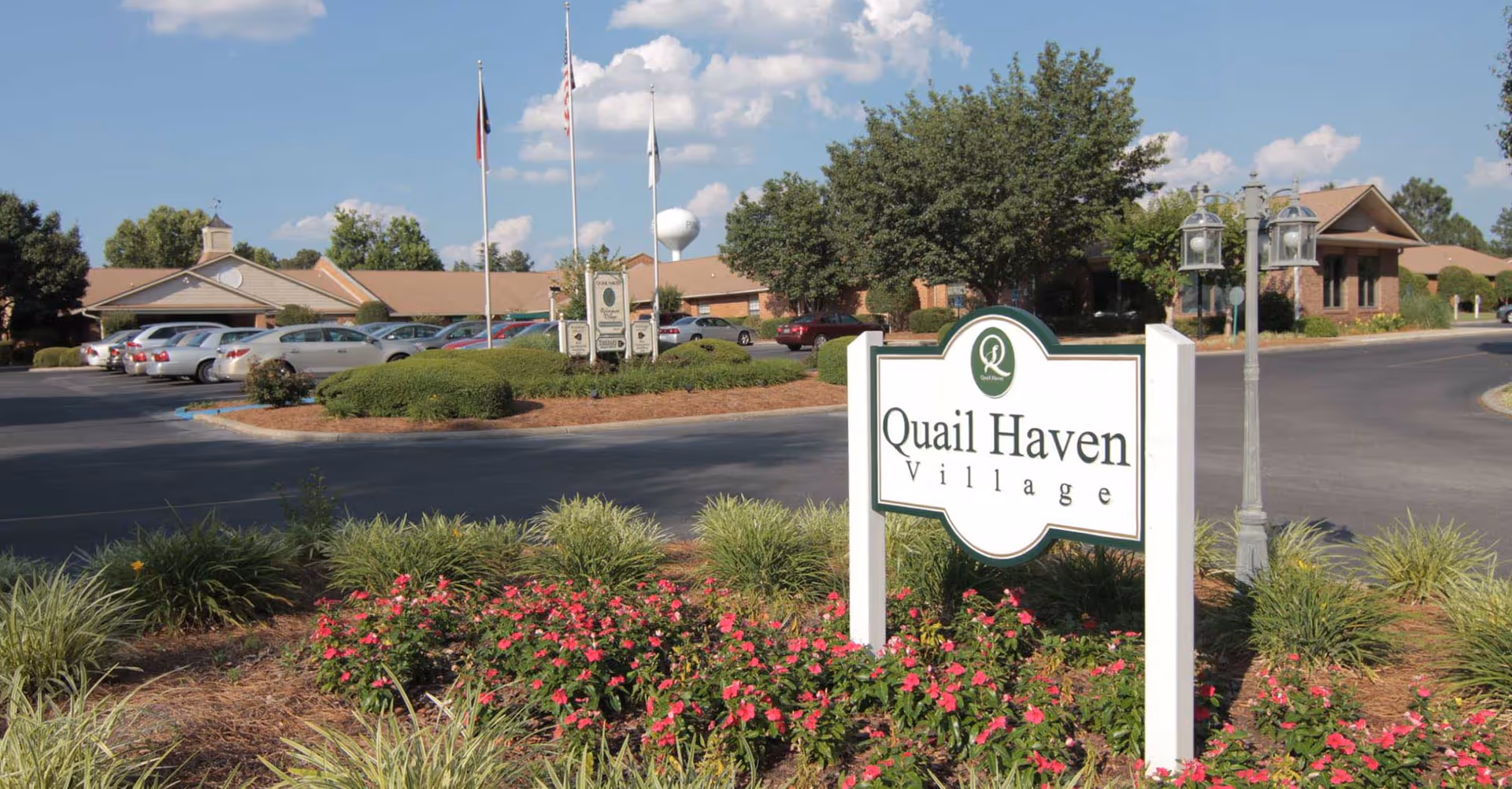 A landscaped entrance sign reading 'Quail Haven Village' with flowers, a driveway, parked cars, and community buildings behind it.