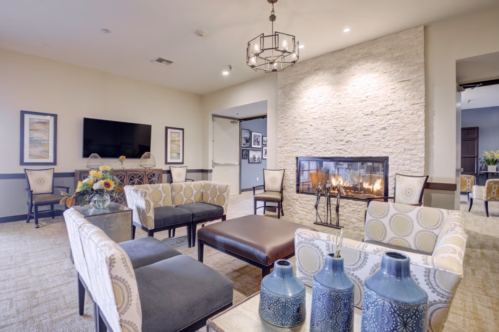 A cozy living room area in an assisted living facility featuring patterned armchairs and sofas arranged around a large brown ottoman. A modern fireplace with a stone surround is lit, providing warmth and ambiance. The room also includes a flat-screen TV mounted on the wall, framed artwork, and decorative vases on a table in the foreground. Soft lighting and neutral tones create a welcoming atmosphere.