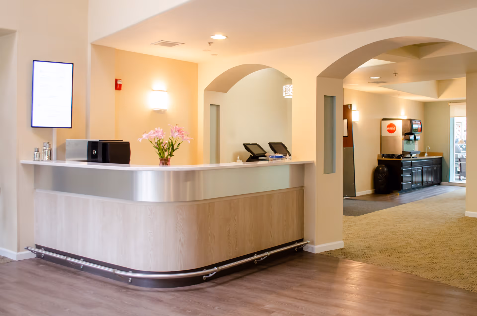 Reception desk and lobby area of a senior living facility with a curved front desk, flowers, and a beverage station in the background.