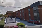 Exterior view of Cliveden Nursing And Rehabilitation Center, a multi-story brick building with several windows. There are cars parked along the driveway and a sign in front displaying the facility's name. The sky is partly cloudy.