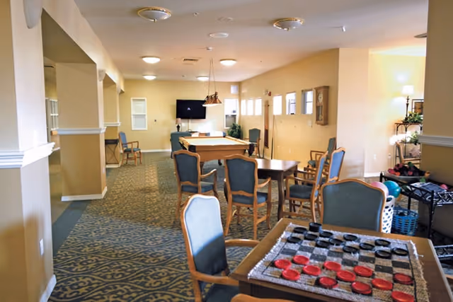 A senior living common room with tables and chairs, a checkers board in the foreground, and a pool table and TV in the background.