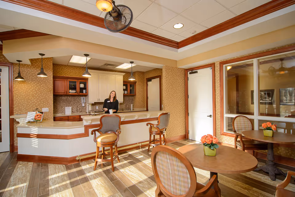A bright and welcoming interior space featuring a reception desk with a smiling woman standing behind it. The room has warm-toned walls, wooden trim, and a ceiling fan with a light fixture. In the foreground, there are round tables with chairs, each table decorated with a small vase of orange flowers. The area appears clean and inviting with natural light streaming in through windows.