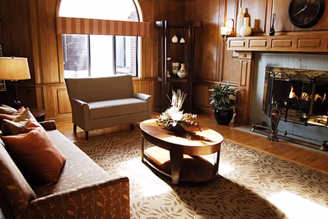 A cozy living room with wood-paneled walls, a large window with a striped valance, a beige loveseat, a patterned sofa with cushions, a wooden coffee table with a floral centerpiece, a fireplace with a fire burning, and decorative items on shelves and mantel.