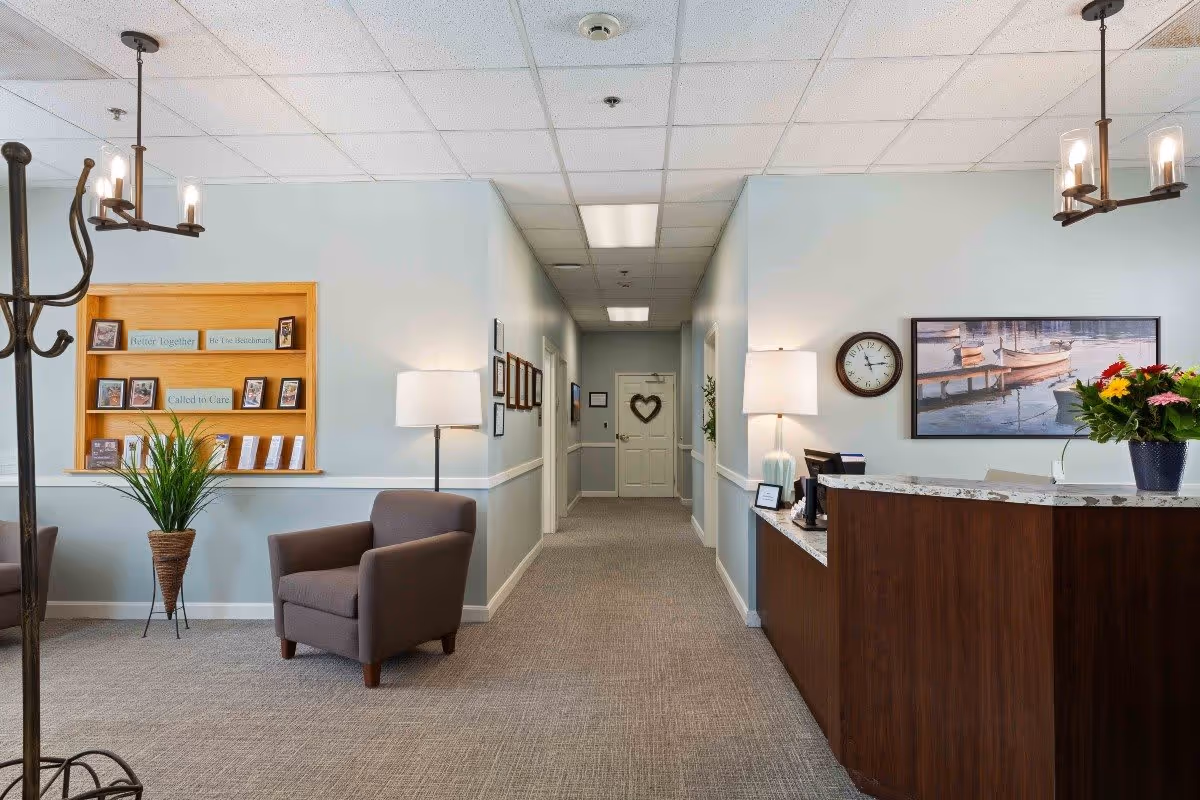Reception area of a senior living facility with a wooden front desk on the right, decorated with a vase of flowers and a lamp. A hallway extends straight ahead with a door at the end featuring a heart decoration. On the left, there is a cozy seating area with two armchairs, a floor lamp, a plant, and a wooden display shelf with framed pictures and signs. The walls are painted light blue with white trim, and the ceiling has recessed lighting and hanging light fixtures.