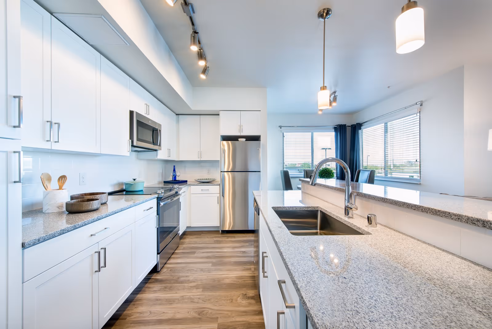 Bright modern kitchen with white cabinets, stainless steel appliances including a refrigerator, oven, and microwave. Granite countertops with a sink and faucet on the island. Wooden floor and large windows with blinds letting in natural light. Pendant lights hang from the ceiling.