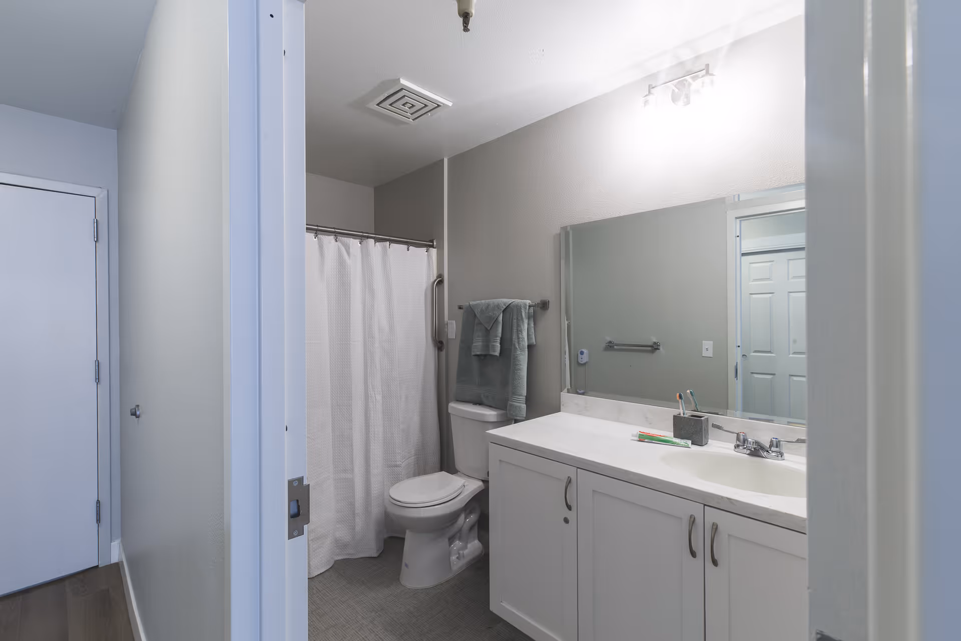 A clean and modern bathroom with a white toilet, a shower with a white curtain, a large mirror above a white vanity with a sink, and two green towels hanging on a towel rack. The walls are painted light gray and the floor has gray tiles.
