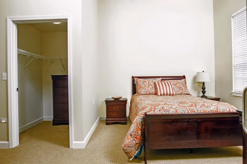 A bedroom with a wooden bed frame and a patterned bedspread with matching pillows. There are two wooden nightstands on either side of the bed, one with a lamp and the other with a small bowl. To the left, there is an open doorway leading to a walk-in closet with a chest of drawers and wire shelving. The room has beige walls and carpeted flooring, with a window on the right side.